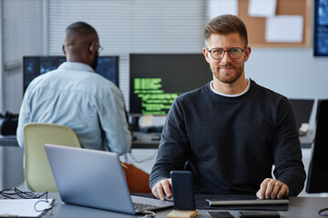 Portrait of male software developer smiling at camera while sitting at workplace in office, copy...