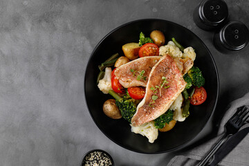 Fillet of fish perch with vegetables, broccoli, cauliflower, tomatoes, potatoes on a dark plate and grey background. Seafood diet plate.