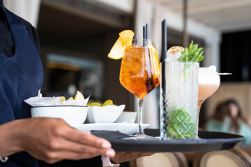 Closeup of tray with cocktails and appetizers - Spritz and mojito served by an African waitress in...