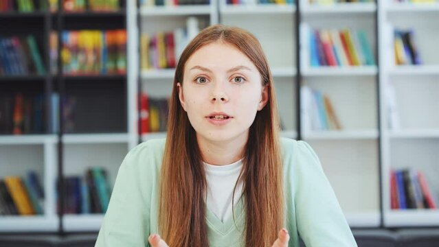 Emotional Young female student with long hair talking to webcam and gesturing during online meeting, blurred bookshelve background. Closeup of young female expressing opinion. Concept of communication