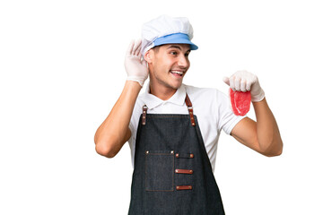 Butcher caucasian man wearing an apron and serving fresh cut meat over isolated background listening to something by putting hand on the ear