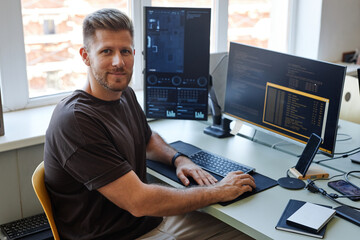 Portrait of young software engineer looking at camera while writing code at workplace with multiple...