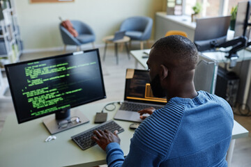 Back view of African American software developer writing code at workplace in office with multiple devices