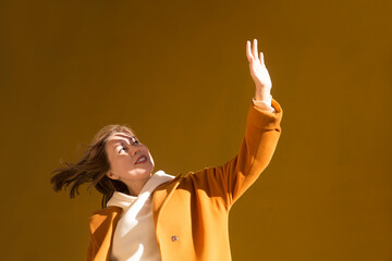 a woman in a yellow coat shades her face with her hand from the sun against the background of a yellow wall walks around the city.  copy space. 