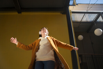 Obraz na płótnie Canvas a woman in a yellow coat with headphones dances and sings on the street of the city against the background of a yellow wall