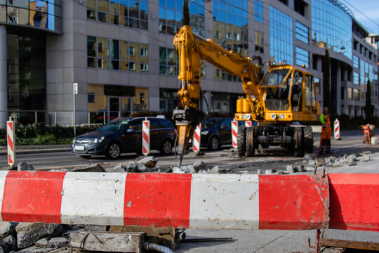 A multifunctional excavator with a hydraulic hammer at the front crushes the asphalt road surface. Red-and-white barrier warning road users about repair work on the road. Opening of asphalt pavements