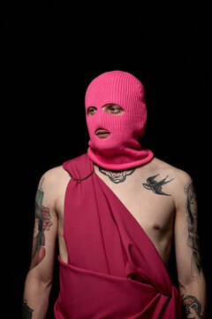 Portrait of young man in pink mask with tattoos on his body standing against black background