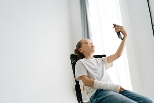 Charming Blonde Little Girl With Broken Arm Wrapped In White Plaster Bandage Talking Video Call On Mobile Phone Sitting In Chair At Light Studio Room. Concept Of Child Insurance And Healthcare