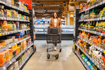 a woman with a cart walks between rows of shelves in a grocery store
