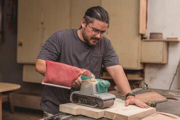 A man uses a belt sander to level the surface of glued wood for a cabinet, table or other furniture at a workshop.
