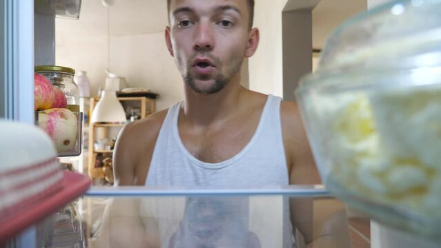 Young guy looking for food at refrigerator to has a breakfast at home. Sleepy man taking oiler and closing fridge at cuisine. Lazy human going to makes brunch at morning. Point of view Slow motion