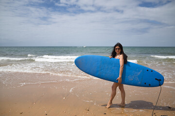 Naklejka na ściany i meble South American woman, young and beautiful, brunette with sunglasses and swimsuit, coming out of the water holding a blue surfboard. Concept sea, sand, sun, beach, vacation, surf, summer.