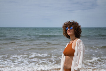 Fototapeta na wymiar Attractive mature woman with curly hair, sunglasses and bikini, posing on the beach happy. Concept sea, sand, sun, beach, vacation, summer.