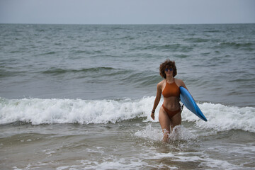 Attractive mature woman with curly hair, sunglasses and bikini, coming out of the water holding a blue surfboard under her arm. Concept sea, sand, sun, beach, vacation, surf, summer.
