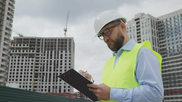 Building inspector wearing helmet and safety vest holding file folder and making notes against multistory buildings under construction. Low angle arc inspector working at site and checking quality