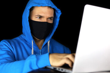 Cybercrime, hacking and technology crime. Man in mask and blue hoodie sitting with laptop on black...