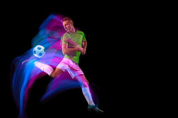 Sport in action. One man professional soccer player training with football ball isolated on dark background in neon mixed light. Sport, speed, power and energy