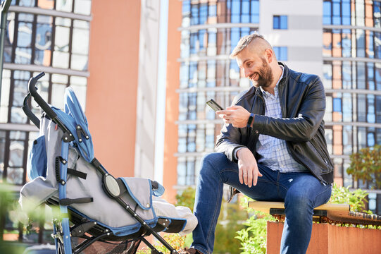 Loving father taking picture of newborn kid, while spending time together outside. Side view of young smiling man taking photo of toddler in pram while sitting on street. Concept of parenthood.
