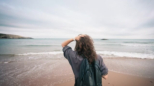 Slow motion backshot of a young caucasian woman with a backpack enjoying the fresh breeze while standing on the beach.