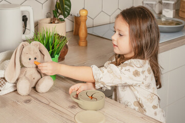 Little dark haired girl 3-4 years old taking piece of carrot out from pastel gray silicone snack cup and feeding toy bunny at wooden table. Baby accessories, tableware, first feeding concept