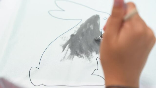 A little girl draws a rabbit with gray acrylic paints, draws on a white T-shirt. The child's hands in close-up work on the fabric. Crafts, preparation for the religious holiday Easter. High quality 4k