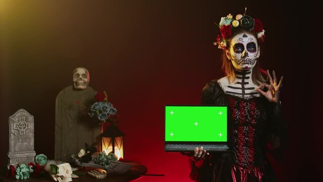 Lady of dead holding laptop with greenscreen, showing isolated copyspace display on pc. Halloween model with creepy body art having isolated chroma key mockup template in studio.
