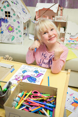 Fototapeta premium A blonde-haired child girl paints a drawing of a unicorn in a children's coloring book. A cheerful child is engaged in fine art.