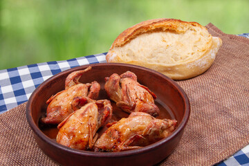 Roasted quail in clay pan and loaf of white bread lie on rustic table with green outdoor background