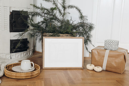 Festive Christmas home decor. Landscape wooden picture frame mockup on parquette floor. Pine, larch tree branches in basket, gift boxes. Cup of coffee. Old white tiled stove background.Empty template