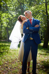 Stylish groom in a blue suit walk with the bride in a white dress in nature. Wedding portrait of happy newlyweds on a background of greenery