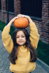 Funny child girl in orange pullover for Halloween with pumpkin and on a dark brick background.