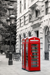Bright red London phone boxes on a city street with lamp post, benches and trees in the background. Black and white with the telephone booths picked out in red