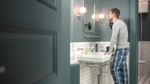 Side view peeping on man brushing his teeth indoor at home. Caucasian handsome young adult male in pajamas standing in bathroom near sink and cleaning teeth with toothbrush looking at mirror