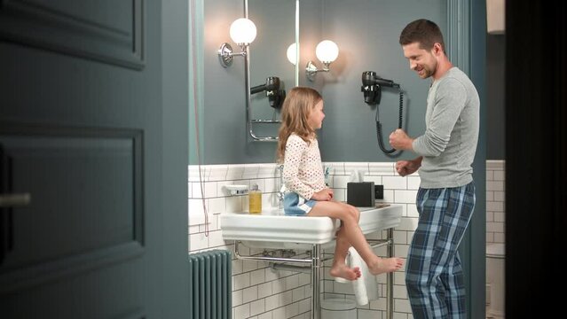 Happy Caucasian handsome father talking with little cute daughter while washing standing in bathroom at home. Young caring dad speaking with child girl in evening. Kid sitting on the sink and smiling