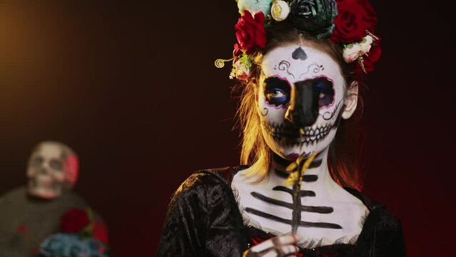 Beautiful model wearing death costume and flowers wreath over studio background, acting like holy santa muerte. La cavalera catrina lady of dead with skull body art, mexican culture. Handheld shot.