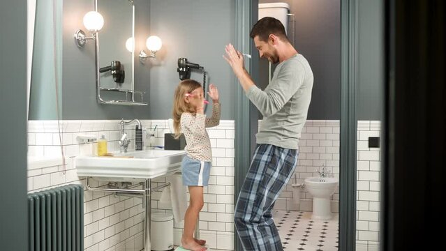 Happy caring young dad and little pretty girl brushing teeth while dancing and singing in good mood. Caucasian caring father with cute small daughter having fun morning dance routine in bathroom