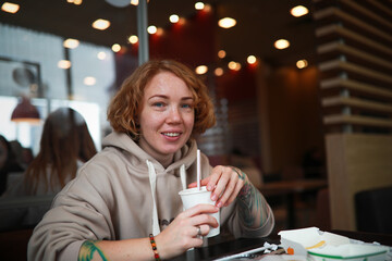 Red-haired young woman with freckle and tattoos drinks juice in cafe with disposable cup through straw