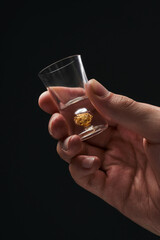 Cropped close-up shot of a man's hand holding an empty shot glass with gold flakes at the bottom. A man holding shot glass with thick base iis on a black background. Front view.