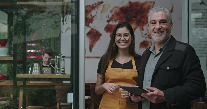 Mature male business manager and young female employee posing together in front of coffee shop small business. Middle aged entrepreneur smiling at camera with barista