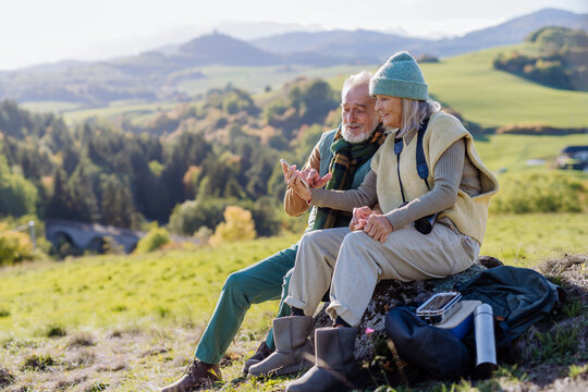 Senior couple having break, looking into phone during hiking in autumn nature.