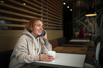 Obraz na płótnie Canvas Woman with smart phone in cafe. Red-haired young woman with freckle and tattoos drinks juice in cafe with disposable cup through straw