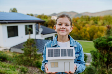Close up of happy little girl holding paper model of house with solar panels.Alternative energy,...