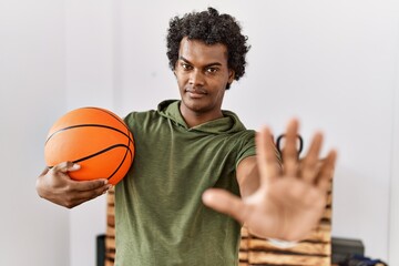 African man with curly hair holding basketball ball at the gym with open hand doing stop sign with serious and confident expression, defense gesture