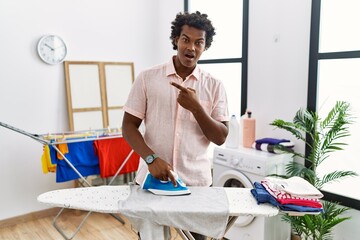 African man with curly hair ironing clothes at home surprised pointing with finger to the side,...