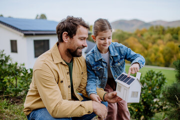 Little girl with her dad holding paper model of house with solar panels, explaining how it...