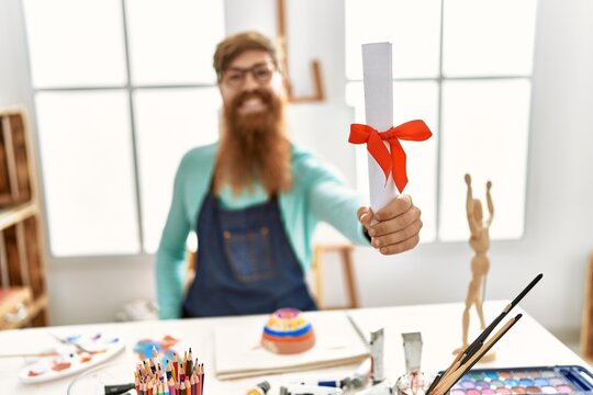 Young redhead man smiling confident holding diploma at art studio