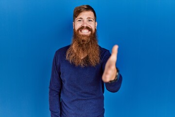 Redhead man with long beard wearing casual blue sweater over blue background smiling friendly offering handshake as greeting and welcoming. successful business.