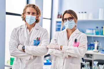 Man and woman wearing scientist uniform and medical mask standing with arms crossed gesture at laboratory