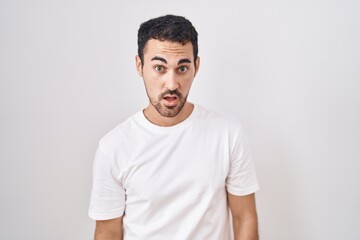 Handsome hispanic man standing over white background in shock face, looking skeptical and sarcastic, surprised with open mouth