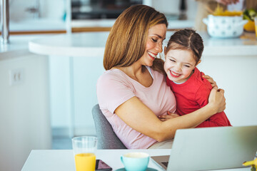 Positive young woman helping daughter in searching information for homework on internet while sitting together at table with laptop at home. Smiling mother and kid having fun